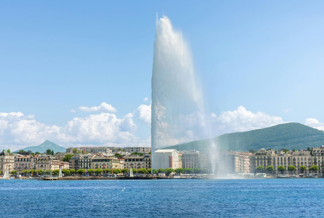 Jet d'Eau fountain on Lake Geneva with Geneva waterfront and mountains under a blue sky