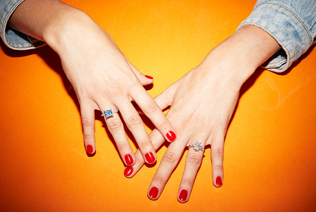 Dos manos con manicura roja muestran anillos de oro con piedra azul y engaste floral de diamantes.