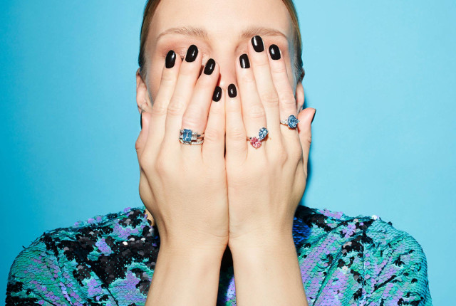 Mani con un anello solitario con diamante blu e un anello con diamante rosa su sfondo azzurro